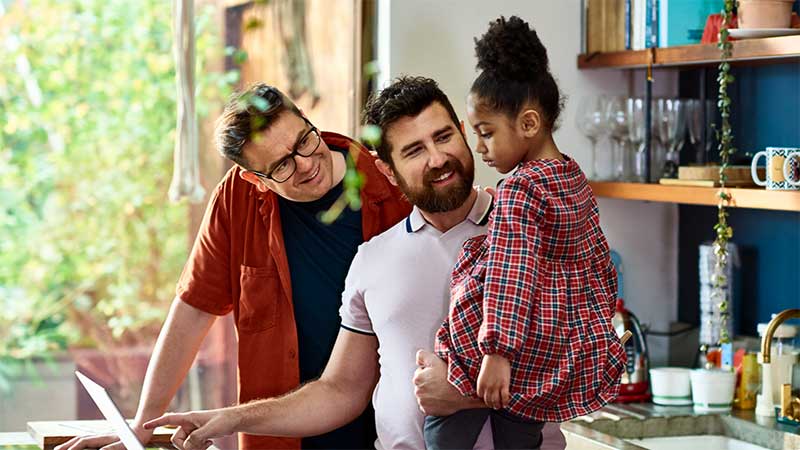 two dads holding their young daughter, and using a laptop