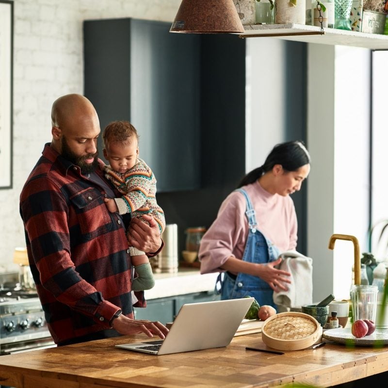 A man working on a laptop while taking care of his young child. In the background is a woman