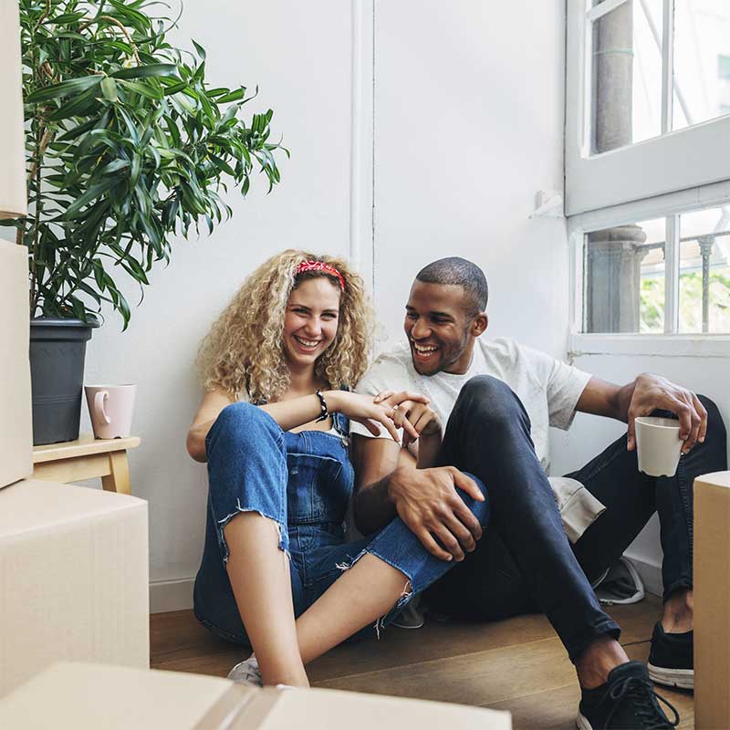 Couple sat on the floor surrounded by moving boxes. They are smiling and laughing