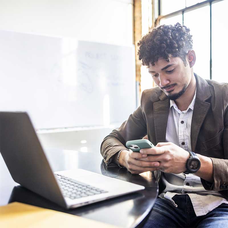 Young man sat a table with a laptop, looking at his phone