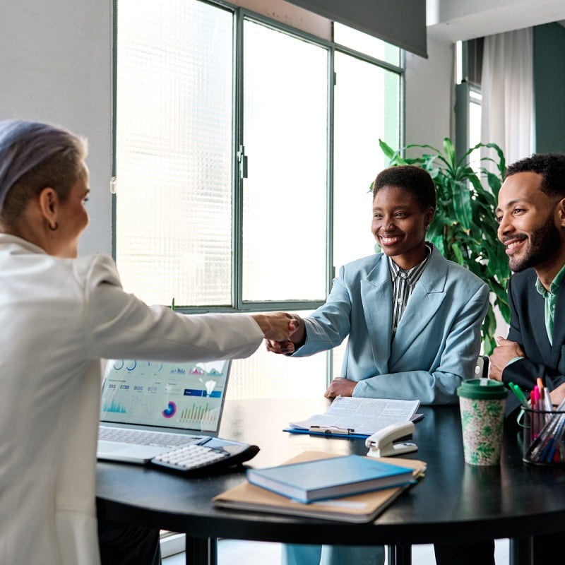 A young couple sat across a desk from an insurance provider. The woman is shaking hands with the provider